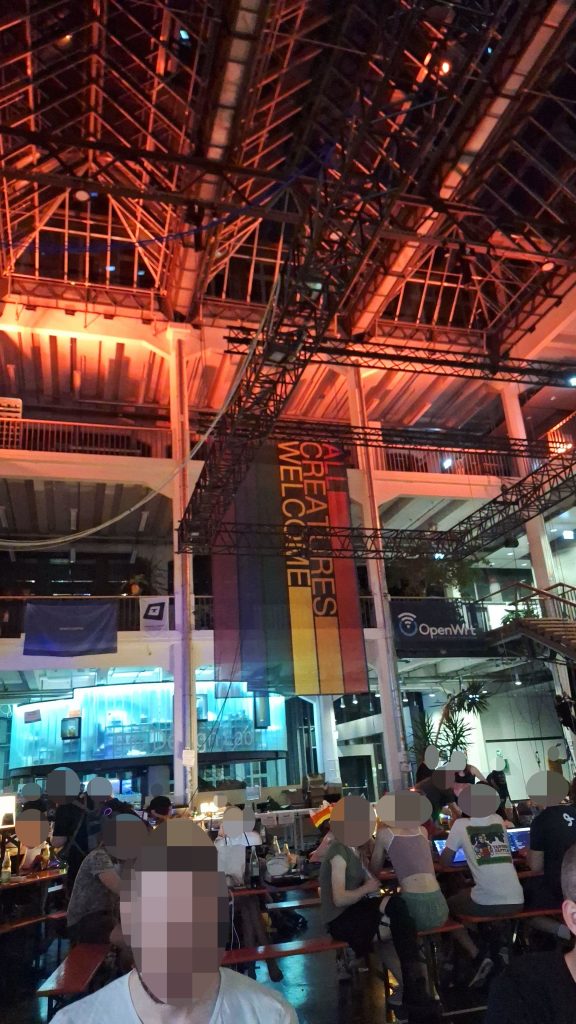 Die Halle der GPN Abends. Die Halle ist indirekt über Led-Streifen beleuchtet, es hängt ein regenbogenfarbenes Banner mit der Aufschrift "All Creatures Welcome" im Zentrum des Bildes. Es sitzen viele Menschen an den Zeltgarnituren und arbeiten an eigenen Projekten. Die Stimmung des Bilds ist angenehm und gemütlich. Die Bildfarben sind vorwiegend warm.